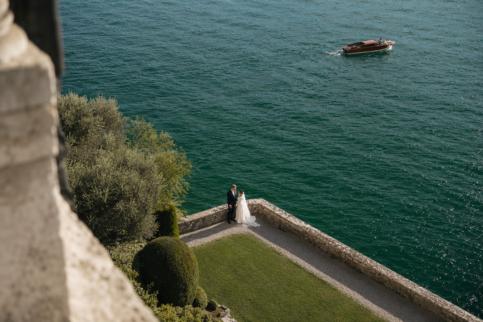 Aerial view of bride and groom at Villa San Vigilio on Lake Garda, Italy, with panoramic lake views and a classic wooden boat.
