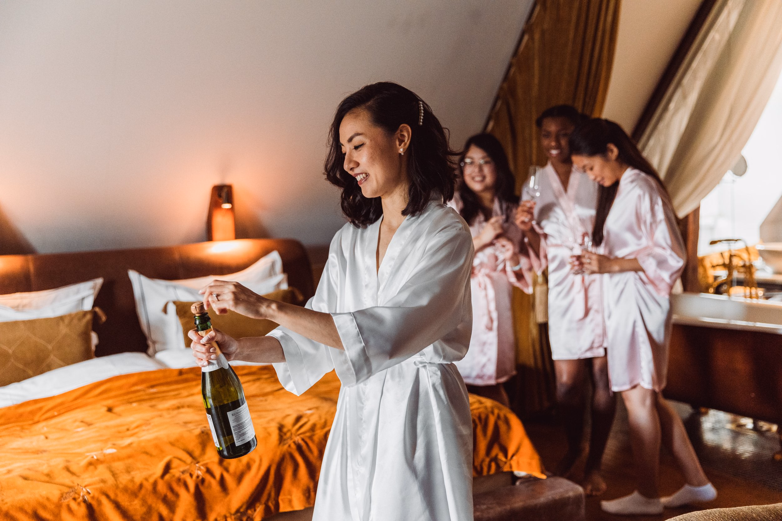 Bride opening a champagne bottle with bridesmaids behind her