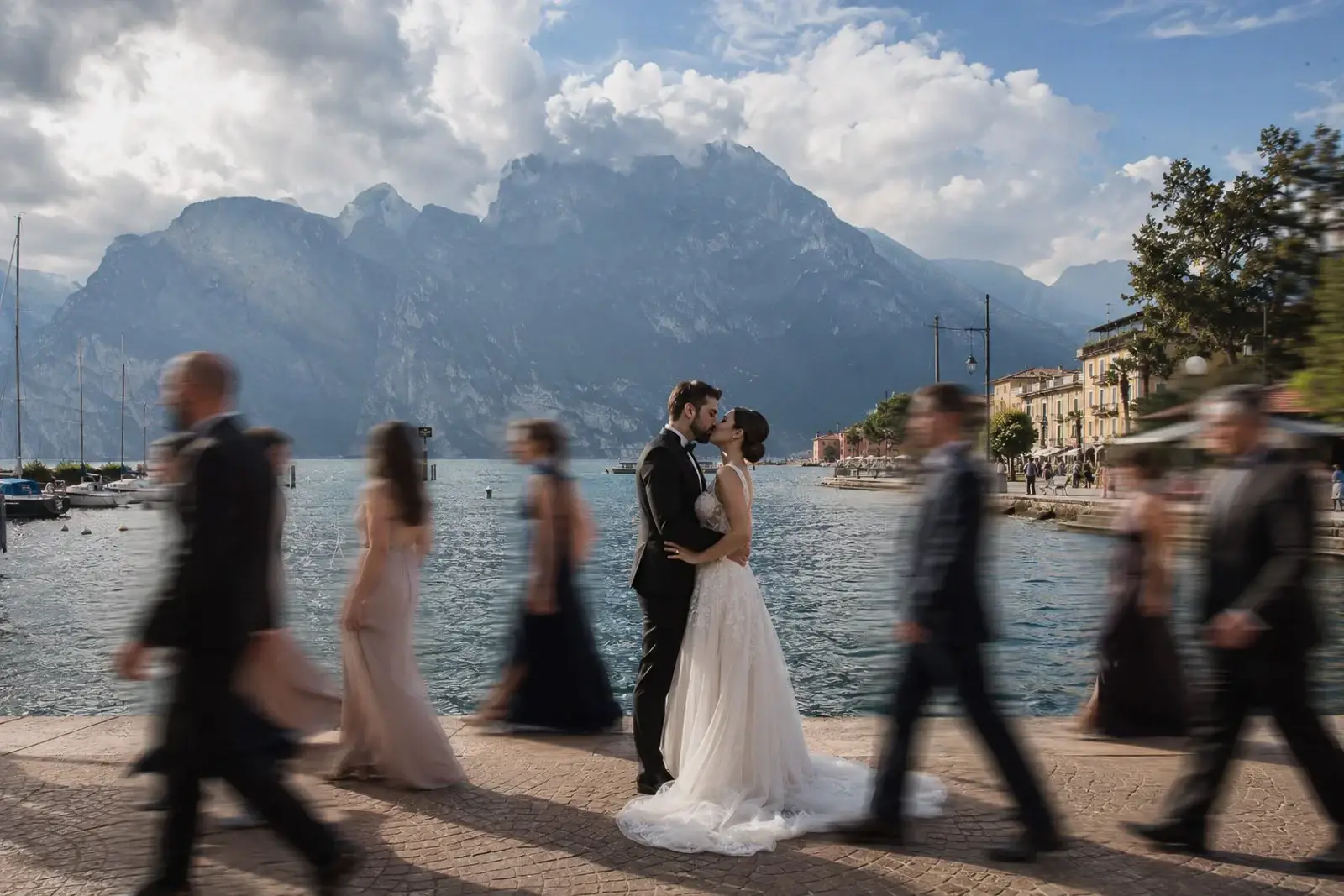 Bride and groom embracing by the waterfront during a destination wedding on Lake Garda, Italy, with dramatic mountain views in the background