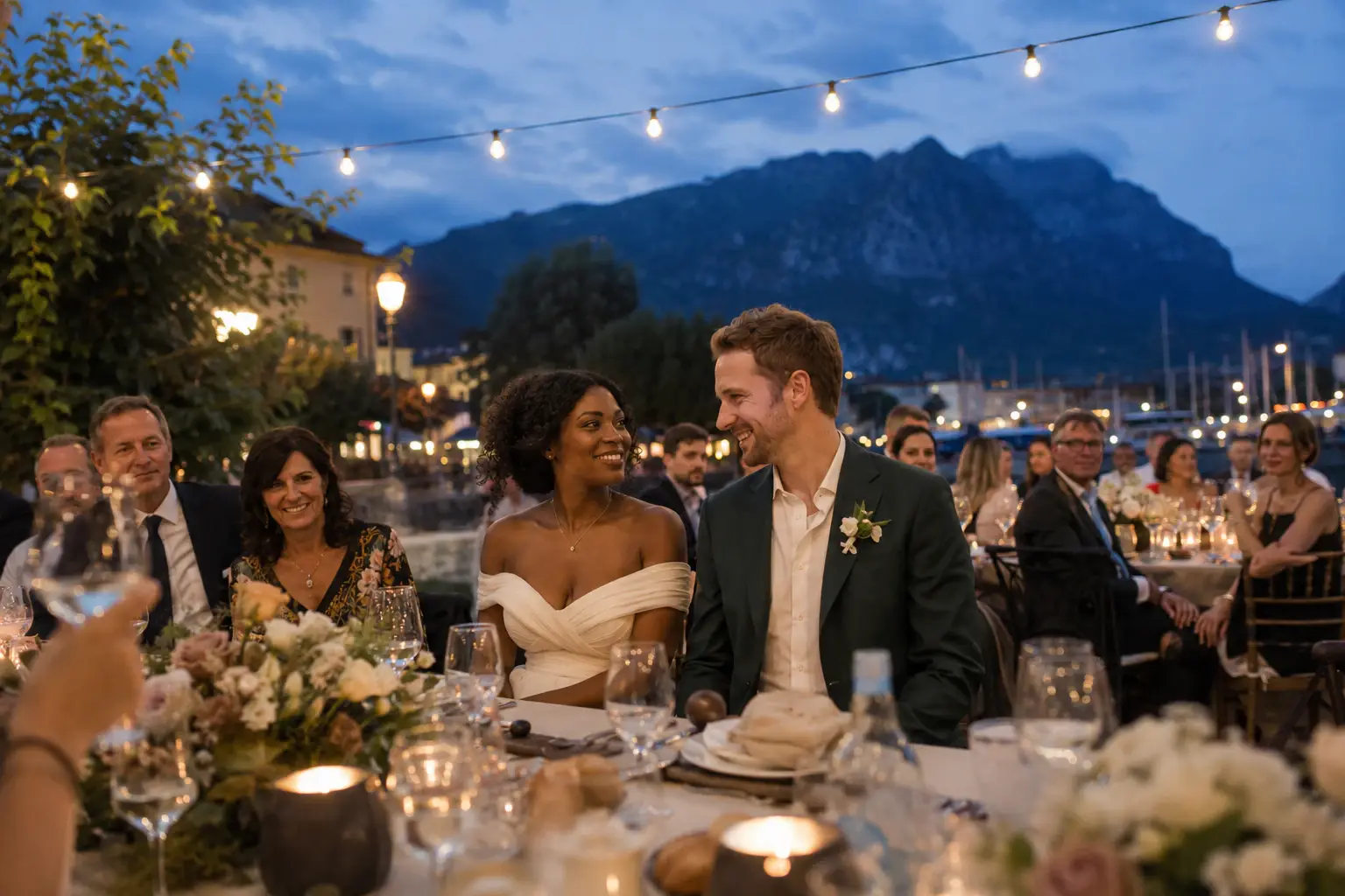 Bride and groom enjoying an outdoor wedding dinner in Riva del Garda with Lake Garda and mountain views at sunset.