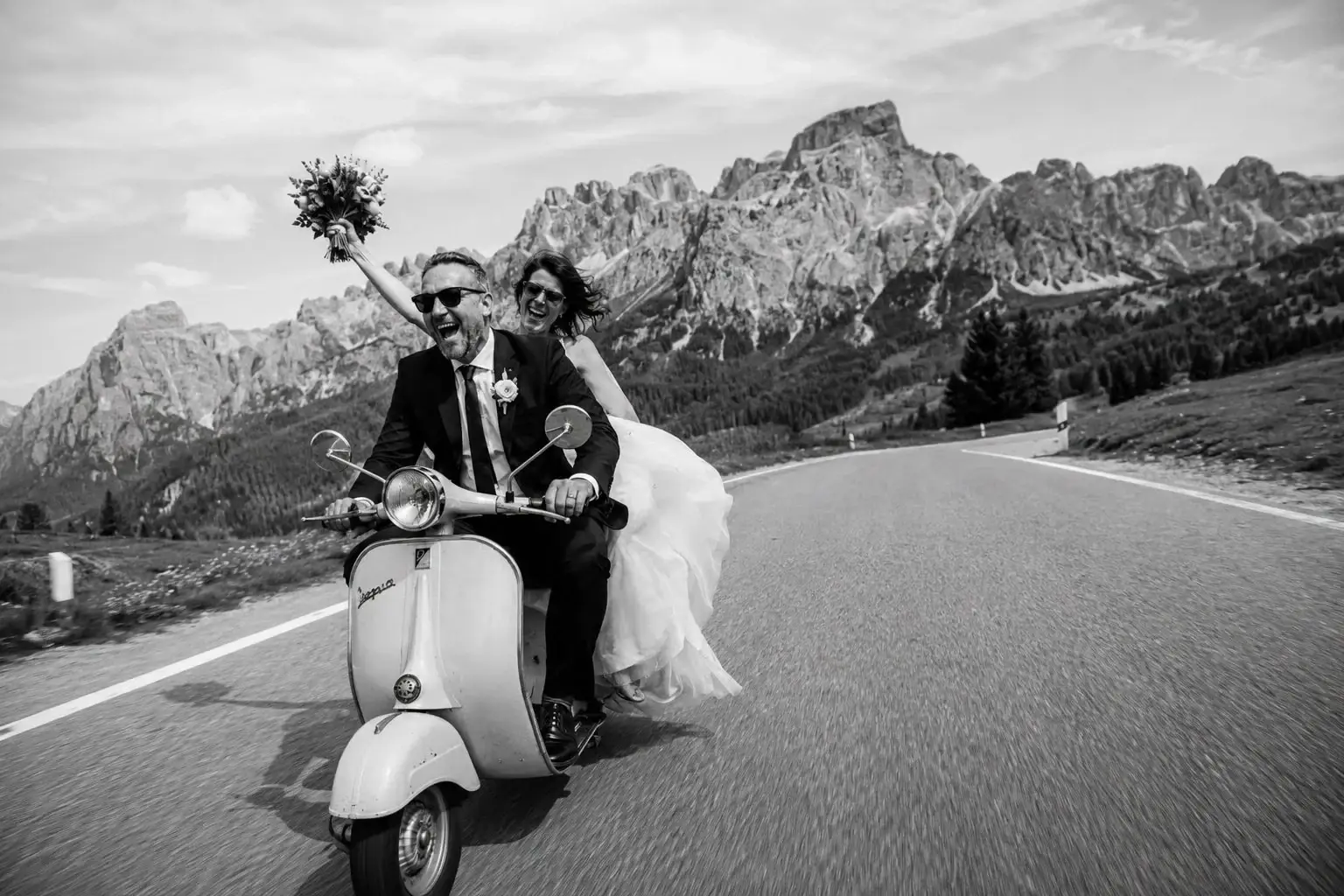 Newlyweds riding a Vespa on a mountain road in the Dolomites.
