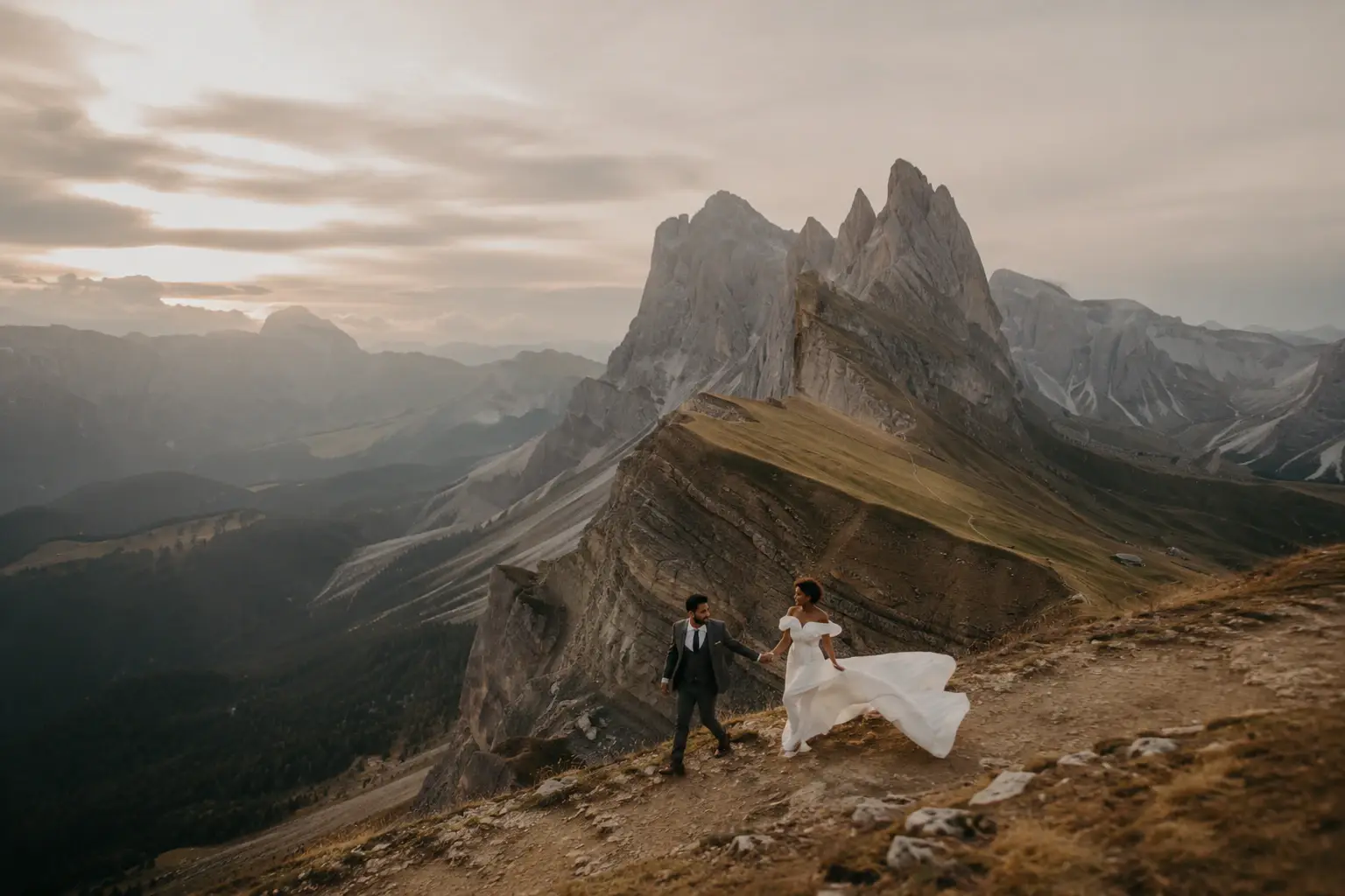 Bride and groom walking along a dramatic mountain ridge at Seceda in the Dolomites during sunset, with sweeping alpine peaks in the background