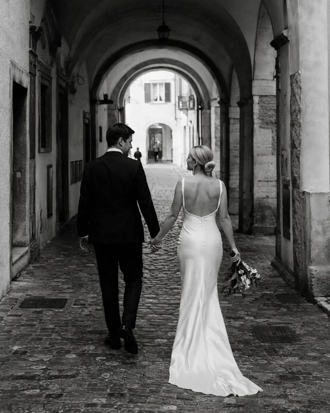 Black and white Trento wedding photo of a bride and groom walking through historic stone arches in Trentino, Northern Italy