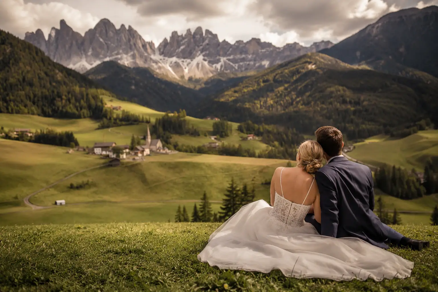 Val di Funes wedding couple sitting on alpine meadow overlooking the Odle peaks in the Dolomites during golden hour.