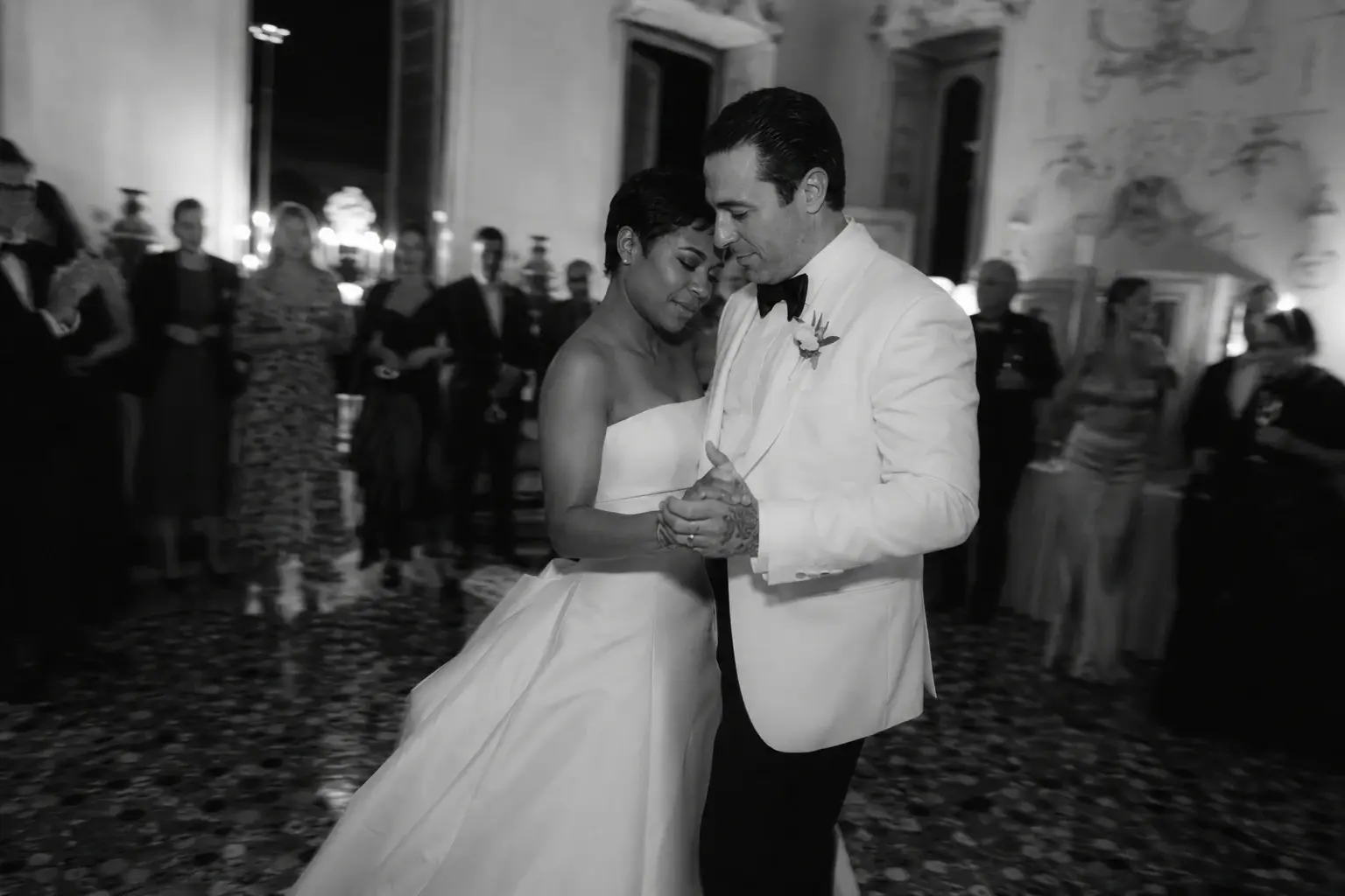 Black and white photo of a couple sharing their first dance inside Villa Sola Cabiati during an intimate Lake Como wedding reception, surrounded by guests and candlelit atmosphere.
