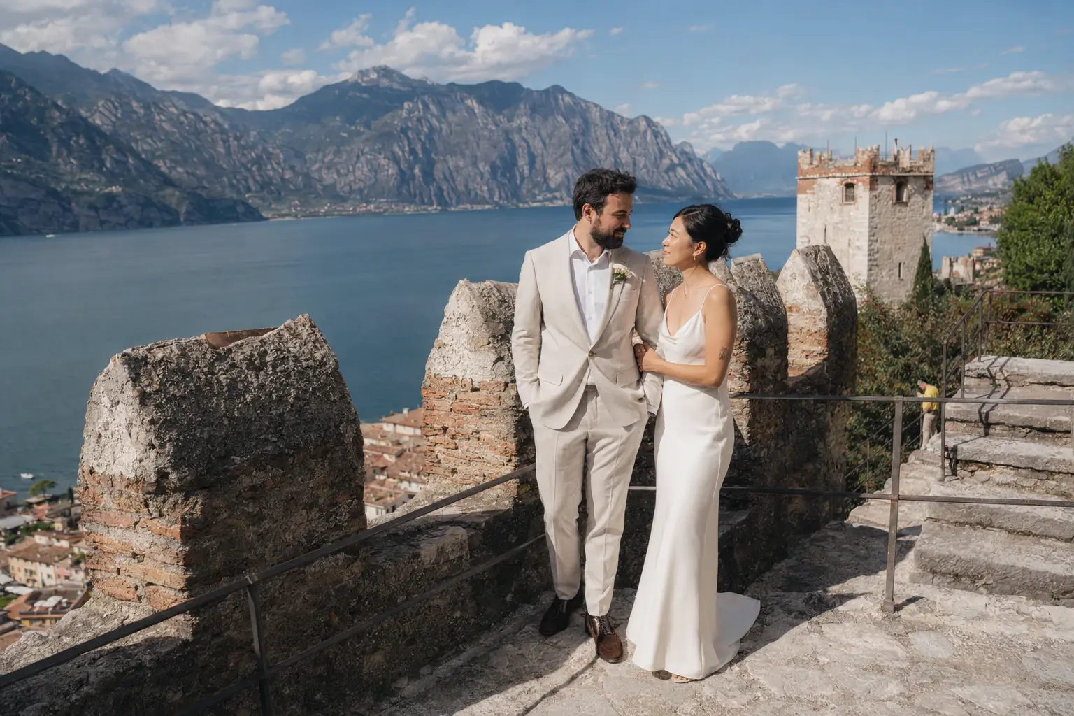 Alt text:  Bride and groom standing on the medieval walls of Castello Scaligero di Malcesine overlooking Lake Garda and the surrounding mountains during a.