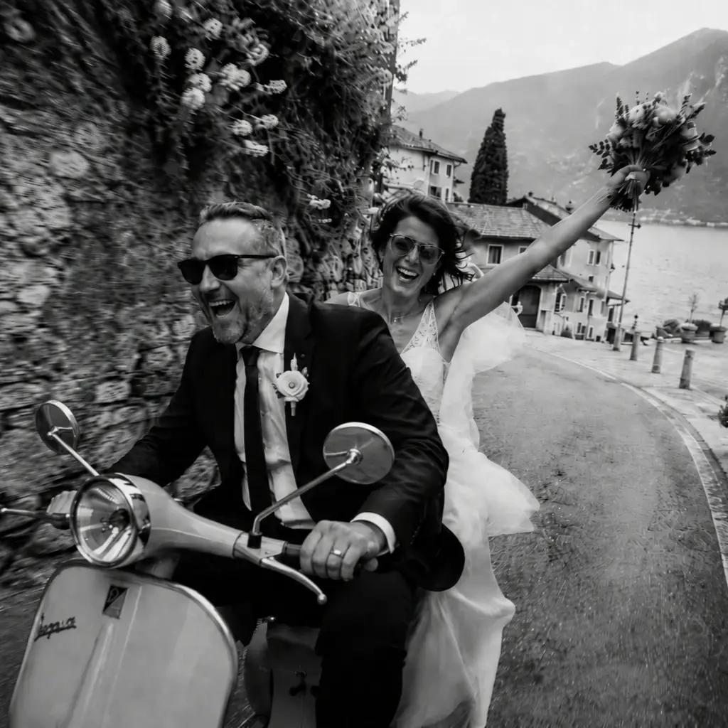 Romantic black and white wedding photo of a bride and groom riding a Vespa through a narrow Italian lakeside village street near Lake Garda.