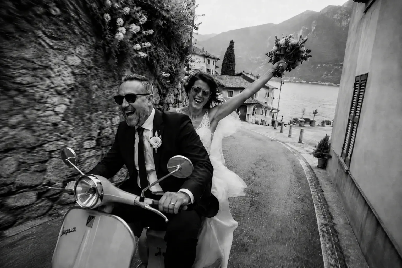 A couple in wedding attire riding a scooter along a scenic street near a lake, with mountains in the background, celebrating and smiling.